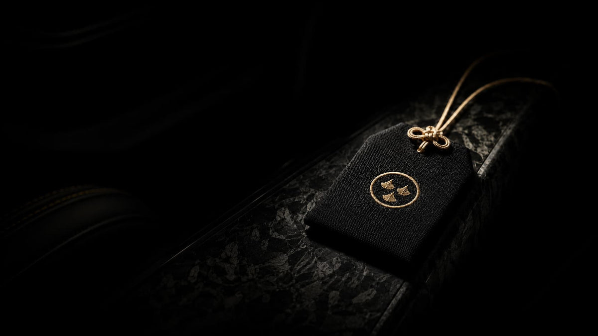 Kirin Scent luxury black omamori car fragrance sachet with gold embroidered logo on a forged carbon fiber console, premium Japanese-inspired automotive interior accessory and luxury gift for car enthusiasts.