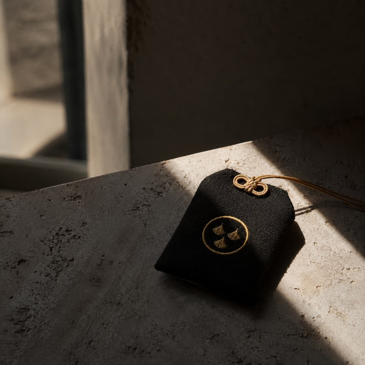 A Kirin Scent 01 Omamori, black fabric pouch with a gold emblem and a gold hanging loop, resting on a textured stone surface in dramatic sunlight and shadow.