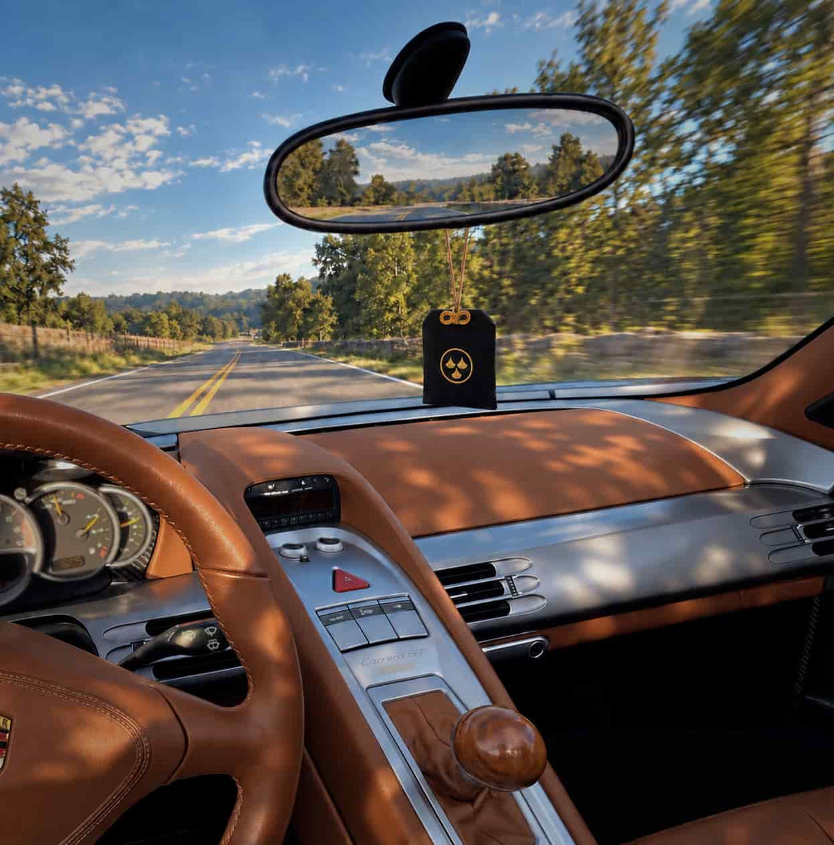 A black car air freshener with a golden Kirin Scent logo hangs from the rearview mirror inside a Porsche Carrera GT's brown leather supercar cabin, with a scenic road and trees visible through the windshield.