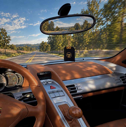 A black car air freshener with a golden Kirin Scent logo hangs from the rearview mirror inside a Porsche Carrera GT's brown leather supercar cabin, with a scenic road and trees visible through the windshield.