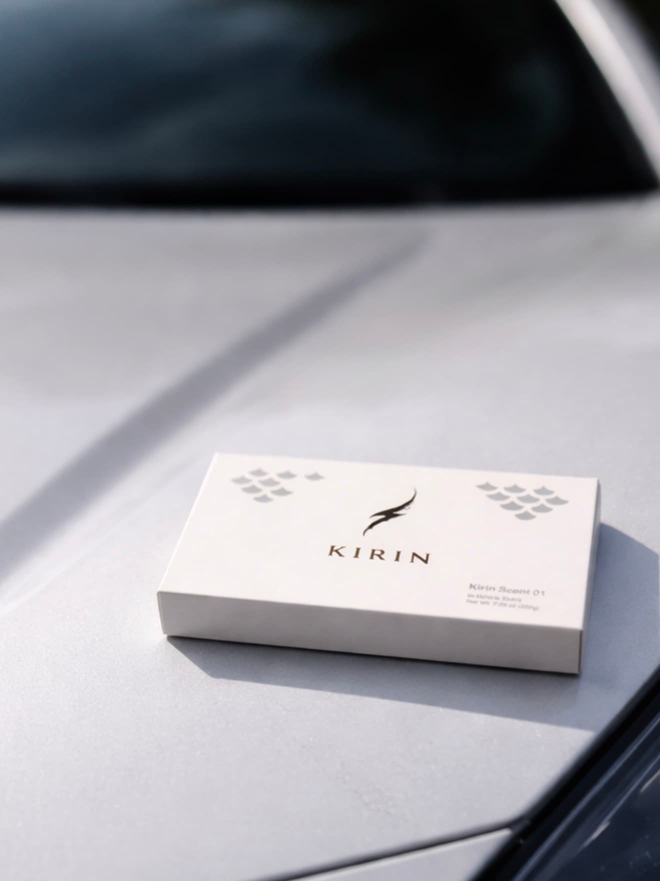 A minimalist Kirin Scent 01 car fragrance gift box placed on the sleek silver hood of a modern luxury car.