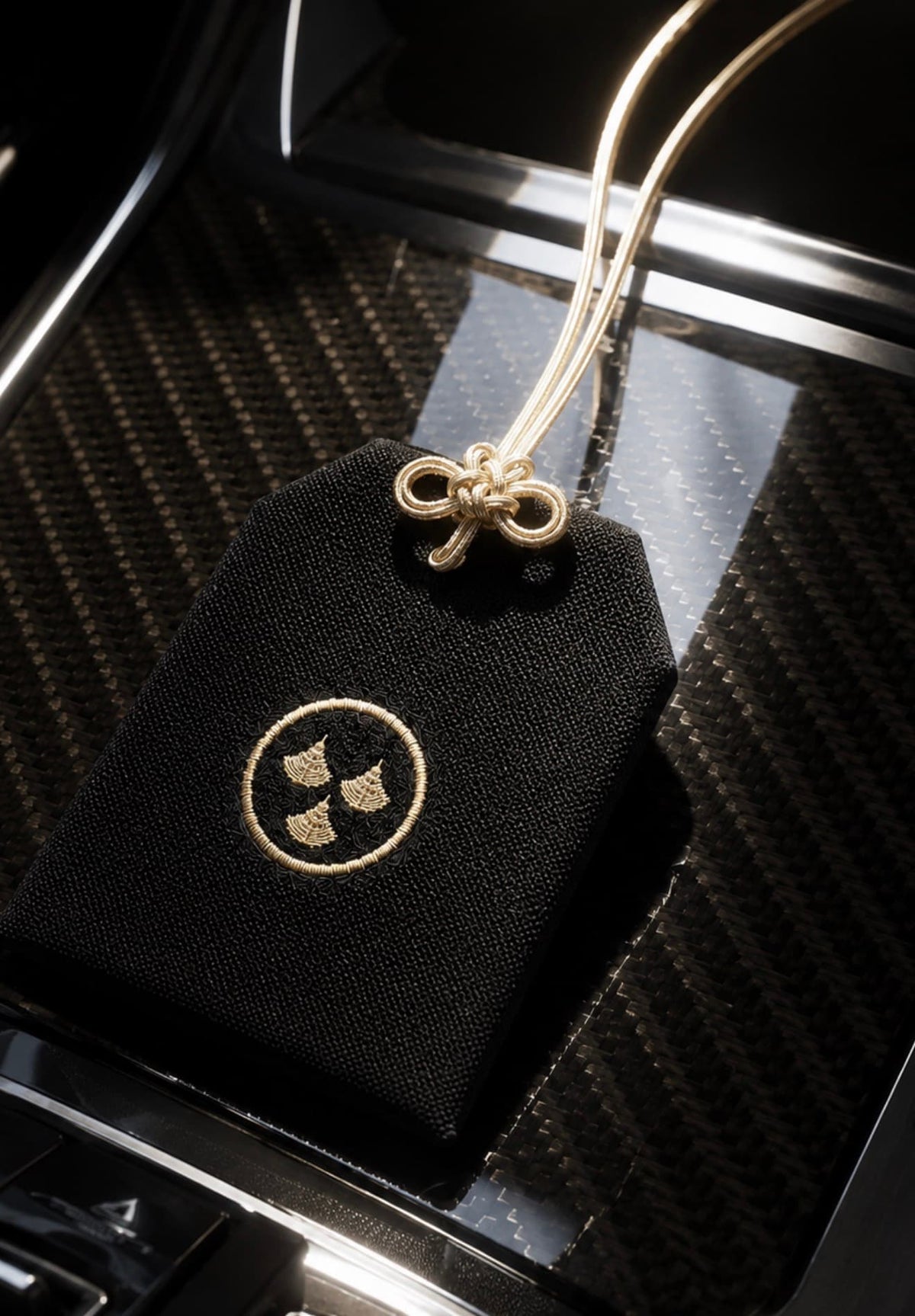 Kirin Scent luxury black omamori car fragrance sachet with golden hanging cords on a glossy carbon fiber interior console, premium Japanese-inspired automotive accessory and gift for car enthusiasts.